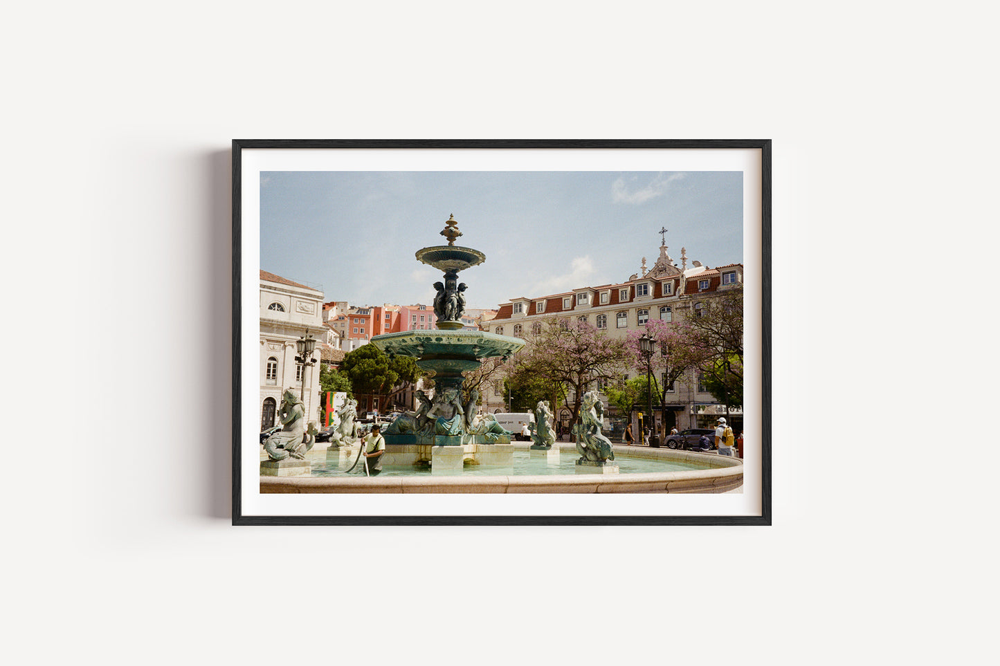 Framed photograph of a fountain in an urban setting with buildings and trees in the background.